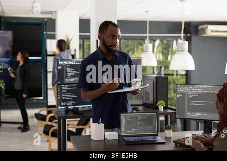 Colleagues discuss technology in office as black man holding clipboard shares updated data for app development. Manager checks progress with employee at computer while working on startup project. Stock Photo