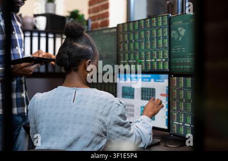 Female stock exchange advisor pointing at computer screen as she explains live updates on investments with her colleague, with black woman seated at desk with multi monitors, analyzing the forex. Stock Photo