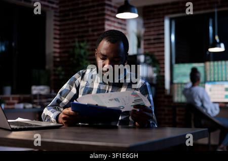 Investment analyst reviewing market documents at night in brick wall office while reviewing charts, market data and financial documents for investment analysis. Stock Photo