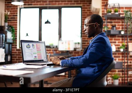 African american man focused on reviewing financial charts displayed on digital device in startup office, connecting office workflow, business planning and professional teamwork. Stock Photo