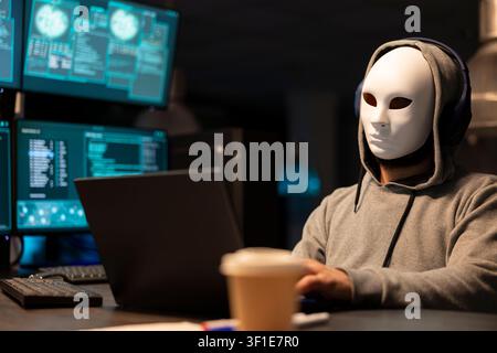 Unidentified man in hoodie wearing white mask as system code displays on screens. Anonymous hacker in office suggests digital secrecy, cybersecurity threats and hidden activity within tech environment. Stock Photo