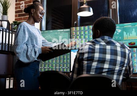 Two financial analysts collaborate at desk in office, reviewing market information on multiple monitors at night, connecting office workflow, business planning and professional teamwork. Stock Photo