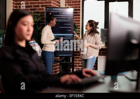 Diverse engineers discuss algorithms on screen, connecting programming code, software teamwork and digital infrastructure during a collaborative IT project. Stock Photo