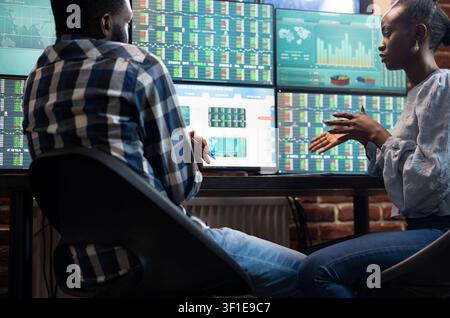 Colleagues analyzing stock charts at desk presenting focused male and female colleagues at desk, analyzing real time stock market charts on multiple monitors in a modern office. Stock Photo