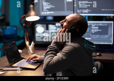 Cheerful male software developer laughing and talking on mobile phone during late night coding session. Caucasian man happily on call, taking short break from programming and decoding office projects. Stock Photo