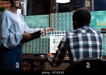 Male and female traders work late in a modern office, reviewing forex charts and stock data across multiple monitors for investment analysis. Stock Photo