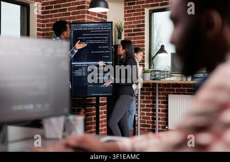 Software developers stand near a vertical screen while discussing an interface bug, slow processing and database security code review, as a software engineering and data workflow scene. Stock Photo