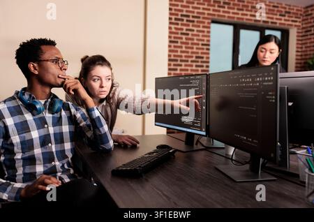 Software developer points to database script on a terminal window while asking a coder for help with HTML code in a data room, with developer teamwork, source code review. Stock Photo