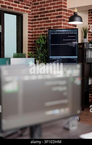 Empty data center office contains multiple computer screens showing programming code, server equipment and modern brick wall technology workspace. Stock Photo