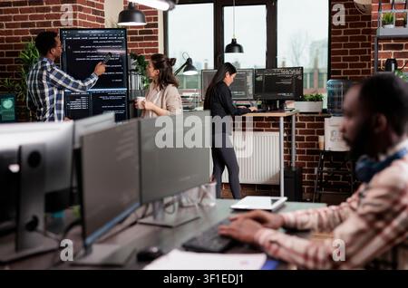 Technology team leader points out a source code mistake while software developers discuss an interface bug and slow data processing near a vertical screen. Stock Photo
