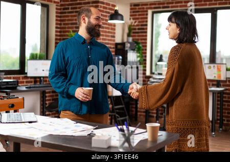 Business partners shaking hands during investment planning as employees use documents, laptops and discussion to manage workplace planning and company decisions. Stock Photo