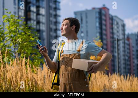 Young male courier holding a cardboard package and checking his smartphone for navigation while standing outdoors in front of residential high-rises, symbolizing fast urban e-commerce delivery Stock Photo