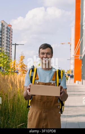 Smiling delivery man courier wearing overalls and a backpack holding a cardboard box package directly towards the viewer, offering fast shipping service in a modern city environment Stock Photo