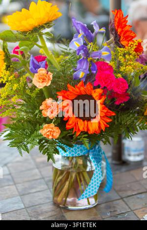A bright, colorful photograph of a diverse floral arrangement featuring yellow and red sunflowers, purple irises, and orange carnations inside a clear Stock Photo