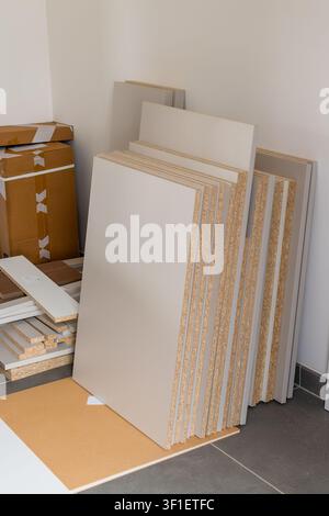 Beige laminate boards stacked vertically against a wall in a spacious interior environment. Various panels and cardboard boxes are visible, indicating preparation for furniture construction. Stock Photo