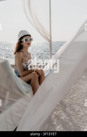 Woman beach hard hat sits in a white cabana by the sea while wearing a safety helmet and sunglasses Stock Photo