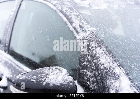 Close-up of a dark car partially covered with fresh snow and frost. The image focuses on the side mirror, window, and roofline, where fine snow crysta Stock Photo