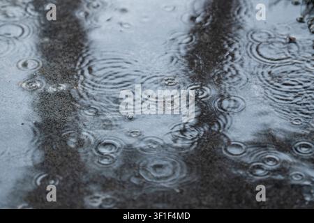 Close-up of raindrops creating circular ripples on wet pavement during rainfall. Abstract rainy weather background with reflections and natural water Stock Photo
