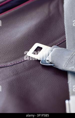A close-up view focusing on the silver metal tongue of an unbuckled grey seatbelt resting against the textured surface of a dark red leather passenger Stock Photo