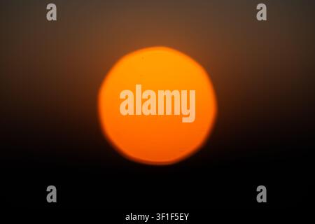 An abstract macro telephoto shot showing a large, vibrant orange sun disk completely blurred out of focus, creating a soft bokeh circle against a dark Stock Photo