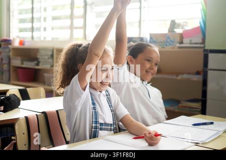 Two cheerful elementary school girls enthusiastically raising their hands while participating in a classroom lesson. Stock Photo