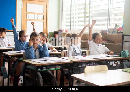 Group of elementary school children sitting at their desks with hands raised while participating in a classroom lesson. Stock Photo