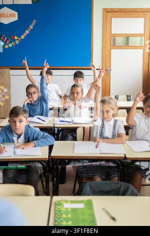Group of elementary school children sitting at their desks with hands raised while participating in a classroom lesson. Stock Photo