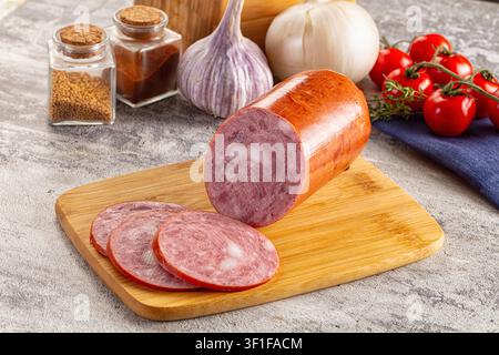 Sliced salami on wooden board with garlic bulbs cherry tomatoes spice jars. Rustic kitchen setup warm lighting natural textures. Ideal for food blogs Stock Photo