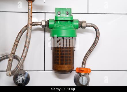 Dirty water filter mesh housing connected with rusty braided hoses on a white tiled wall backdrop. Stock Photo