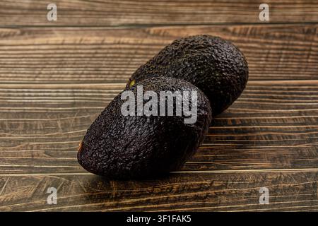 Two dark ripe avocados resting on textured wooden table. Warm lighting highlights natural texture and deep green black skin. Rustic composition ideal Stock Photo
