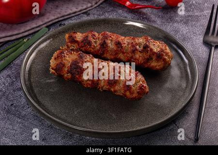 Two grilled meat sausages on dark textured plate. Green onions red pepper in background. Warm lighting highlights charred surface. Rustic food styling Stock Photo