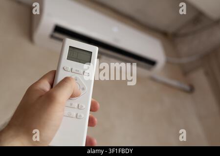 Hand holding a white remote controller to adjust an air conditioner on the wall during hot summer weather. Stock Photo