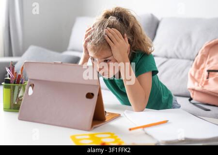 Elementary school girl holding her head while looking at a tablet during homework at home. Stock Photo