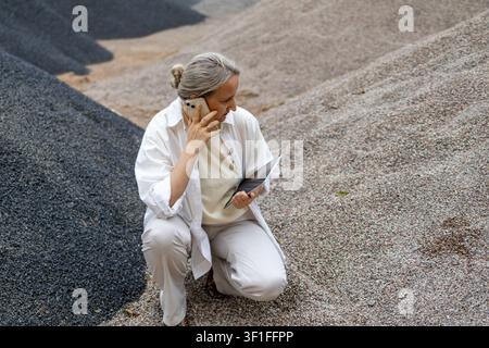Middle aged woman speaks on smartphone while reviewing information on a tablet beside large aggregate stockpile outdoor. Concept of remote consulting, Stock Photo