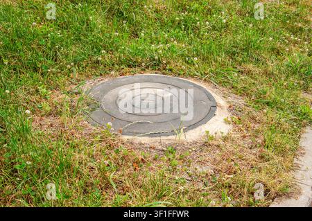 A circular cast iron access point is partially obscured by lush green grass and small white clover blossoms, suggesting an outdoor utility infrastruct Stock Photo