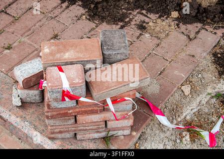 A pile of red and gray interlocking paving stones sits on a brick pathway, marked with red and white caution tape, indicating ongoing construction or Stock Photo