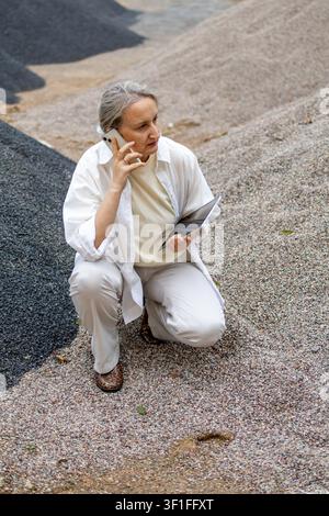 Middle aged woman talks on smartphone while inspecting a construction aggregate stockpile and holding a tablet outdoors. Concept of field communicatio Stock Photo