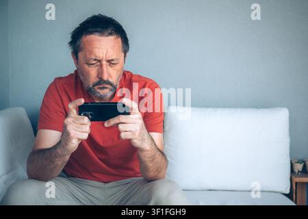 Middle-aged man sitting on sofa focused on smartphone screen, mobile technology and digital communication concept, selective focus Stock Photo