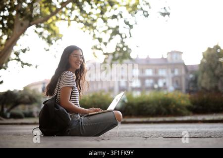 Happy student sitting outdoors using her laptop Stock Photo