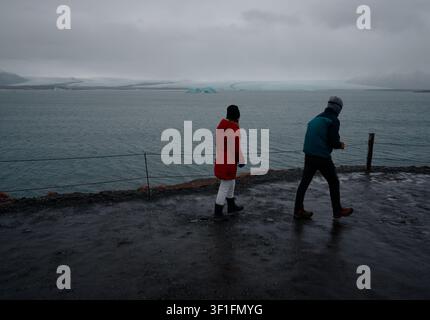 Two visitors walk the rain-soaked shoreline path of Jökulsárlón glacier lagoon, drifting icebergs in the distance Stock Photo