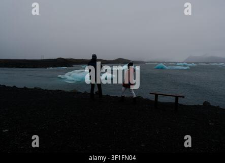 Two visitors walk the shoreline path of Jökulsárlón glacier lagoon with blue icebergs Stock Photo