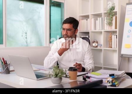 Thoughtful Indian businessman analyzing financial reports on laptop in office Stock Photo