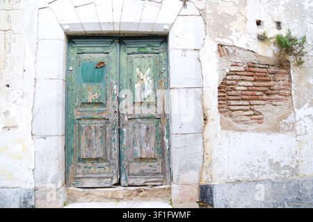 An old weathered green wooden door with peeling paint set in a stone archway on a distressed white plaster wall showing exposed red bricks in the hist Stock Photo