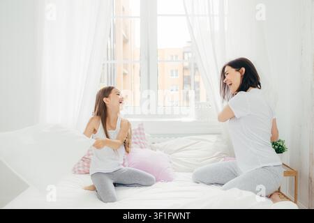 Mother and daughter laughing during pillow fight on bed in bright white bedroom Stock Photo