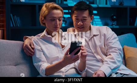 Multiethnic couple emotional discussing shopping using mobile phone at evening living room at couch. Multiracial family Asian man with Caucasian woman Stock Photo