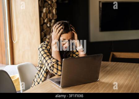 Overwhelmed woman holding her head while working on laptop at home, remote work burnout, digital fatigue, cognitive overload, productivity pressure Stock Photo