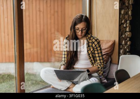 Woman working on laptop by large window at home, calm remote work lifestyle with deep focus, flexible schedule, digital nomad routine, mindful Stock Photo