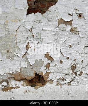 Decaying painted white exterior wall surface with peeling cracked paint, broken concrete layers and holes revealing red brick and crumbling mortar Stock Photo