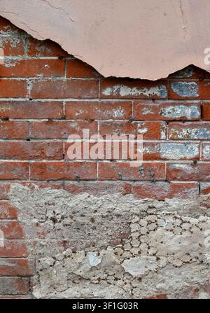 Tan painted wall with cracked paint, deteriorating concrete, exposed rusted metal lath, and worn red brick with crumbling mortar Stock Photo