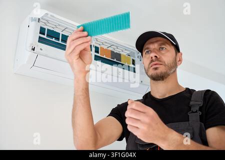 Man checks a removable filter from an open wall mounted air conditioner during cleaning and maintenance at home. Indoor cooling equipment is inspected before servicing. Stock Photo